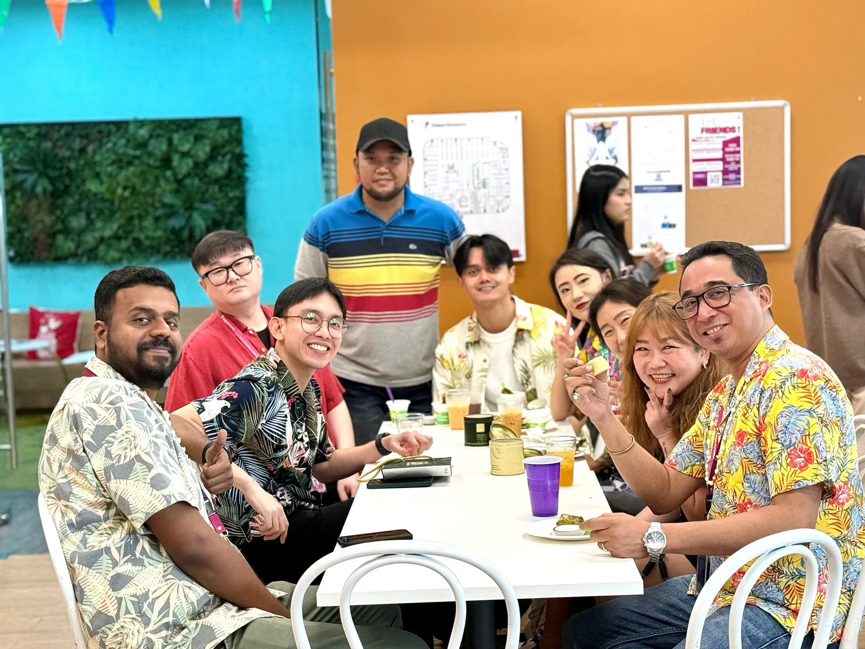 A diverse group of colleagues in vibrant tropical shirts enjoy a cheerful office gathering, sharing food and laughter together